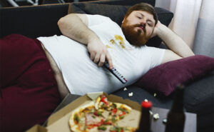 homem obeso deitado no sofá assistindo TV e comendo muitas comidas calóricas e gordurosas